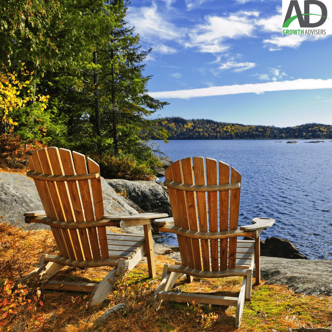 Two wooden Adirondack chairs facing a lake surrounded by autumn trees